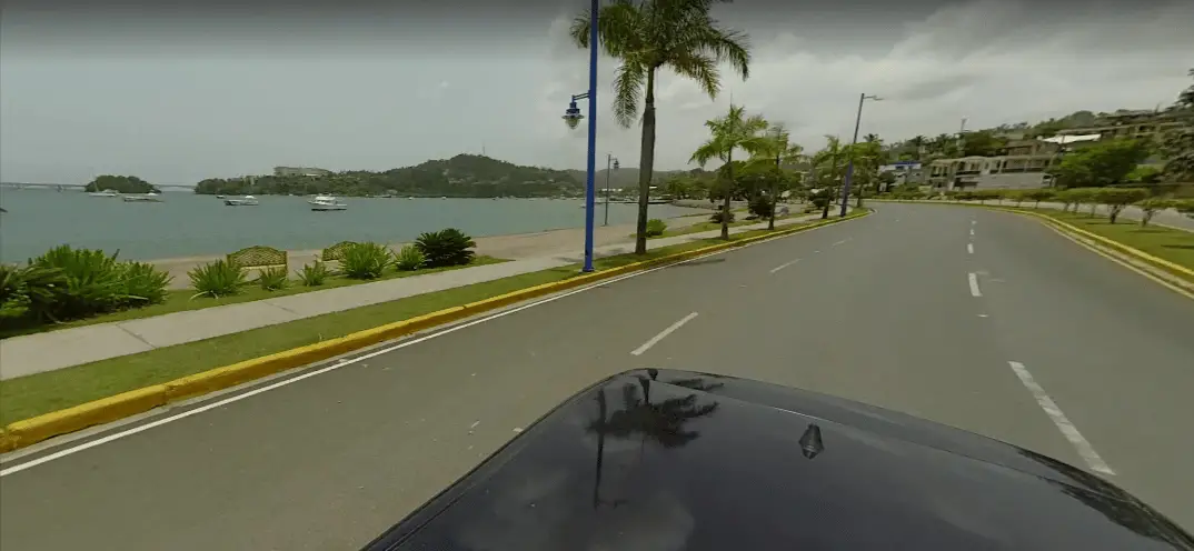 Snapshot from Google Street View showing flat, paved sidewalk between the road and the shore, with palm trees and benches lining the sidewalk. The buildings of the city center are seen in the distance.