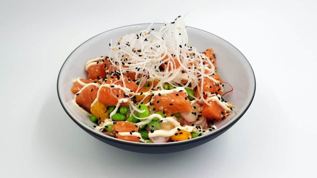 Poke Bowl with rice on bottom, edemame, raw salmon chunks, black sesame seeds, crunchy rice noodles piled on top of the salmon, drizzled with a white sauce