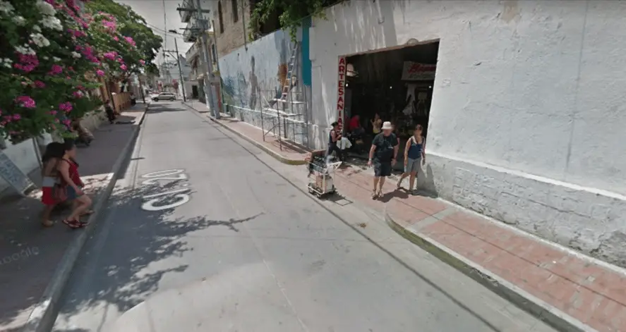 Paved road with sidewalks. Building painted white with garage-sized hole cut in wall which is open for customers to enter. In the shadows inside you can see a sign hanging that says "Bienveido" and products stacked for sale. 