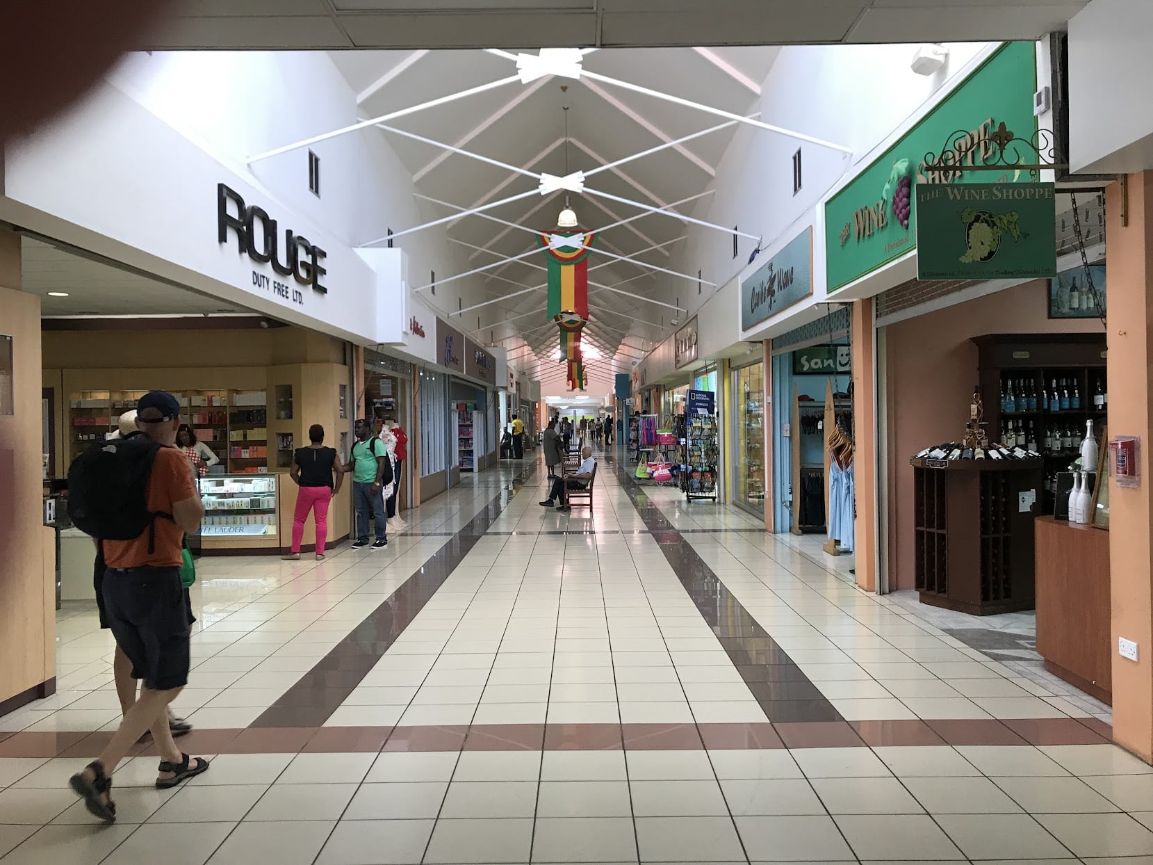 Interior of an indoor shopping mall. A long tiled hallway lined with stores. There are a few people walking down the hall.