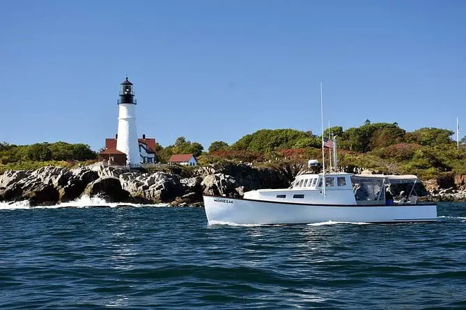 Small white lobster boat sails through dark blue waters past a tall white lighthouse on a rugged coastline.