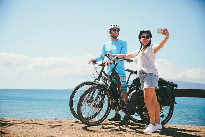 Woman taking a selfie with a man standing behind her. They are straddling e-bikes and are overlooking the ocean.