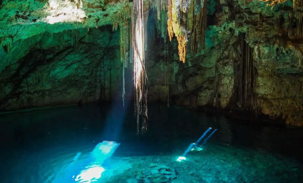 Underground swimming hole with sun rays shining down on the clear blue water. Stalactites hang from the ceiling.