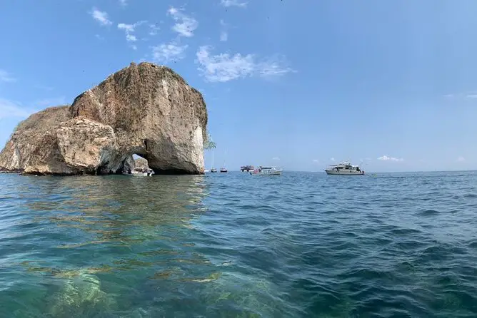 Large arc-shaped rock jutting out of the water, with small boats floating nearby.
