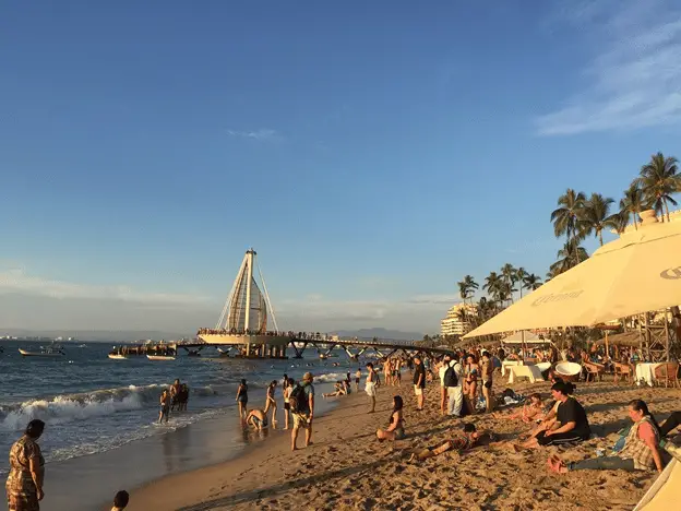 Beach with people walking and lounging on the sand, and people walking and swimming in the water. In the background is the Playa de los Muertos pier with its large sail-shaped metal