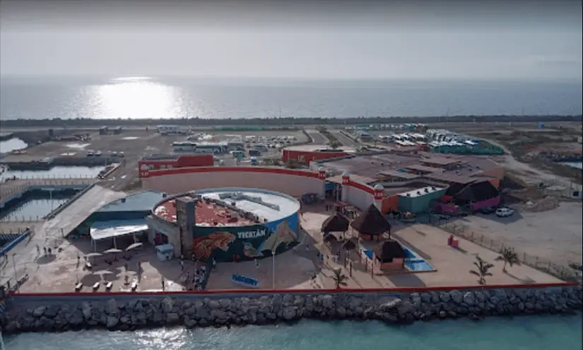 Cruise ship terminal view form the ship. The strip of land is narrow with water visible on both sides. A round building painted colorfully is in the center with a few tables and umbrellas, indicating an eating area. A larger building is directly behind, with access to the road leading down the pier to the shore. 