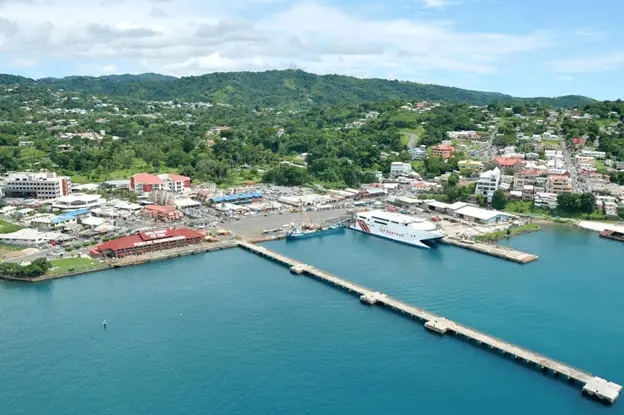 View of pier and port from a ship coming in to dock. Long empty pier surrounded by calm blue water. Pier leads to cruise terminal, surrounded by short buildings. Background is tree-covered mountain with homes and buildings dotting its sides.