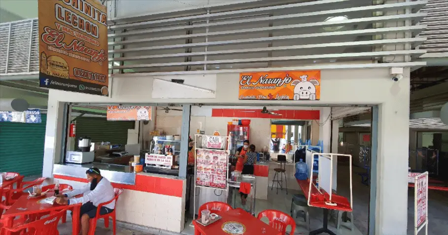 Inside of El Naranjo in the market showing a few red plastic tables and chairs in the foreground with a booth for ordering with an open kitchen in the background. 