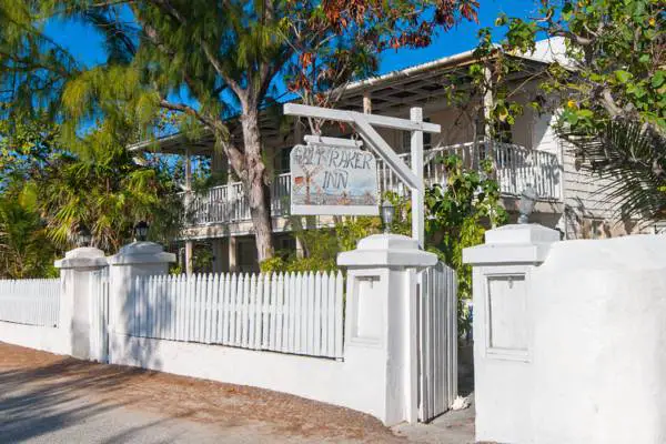 Outside entrance to The Salt Raker Inn as shown by a white sign hanging from a white post. There are cement pillars showing the entrance with a white picket fence on either side. Behind are large leafy green trees providing shade to the full-length balconies of the white two-storey inn.
