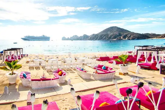 Beach lined with bright pink lounge chairs with a mountain rising up out of the turquoise ocean in the background, with a cruise ship docked beside.