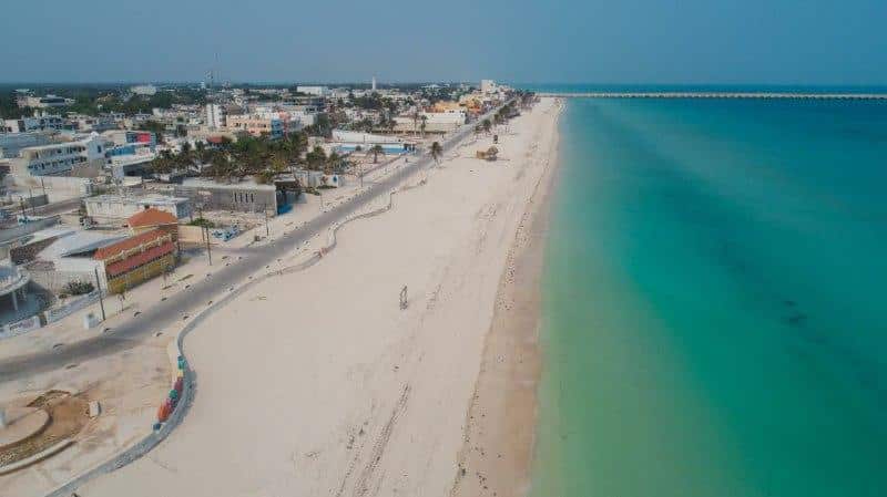Aerial view of wide white-sand beach with greenish-blue ocean on the right side and paved roadway on the left, with buildings sitting along the roadway.