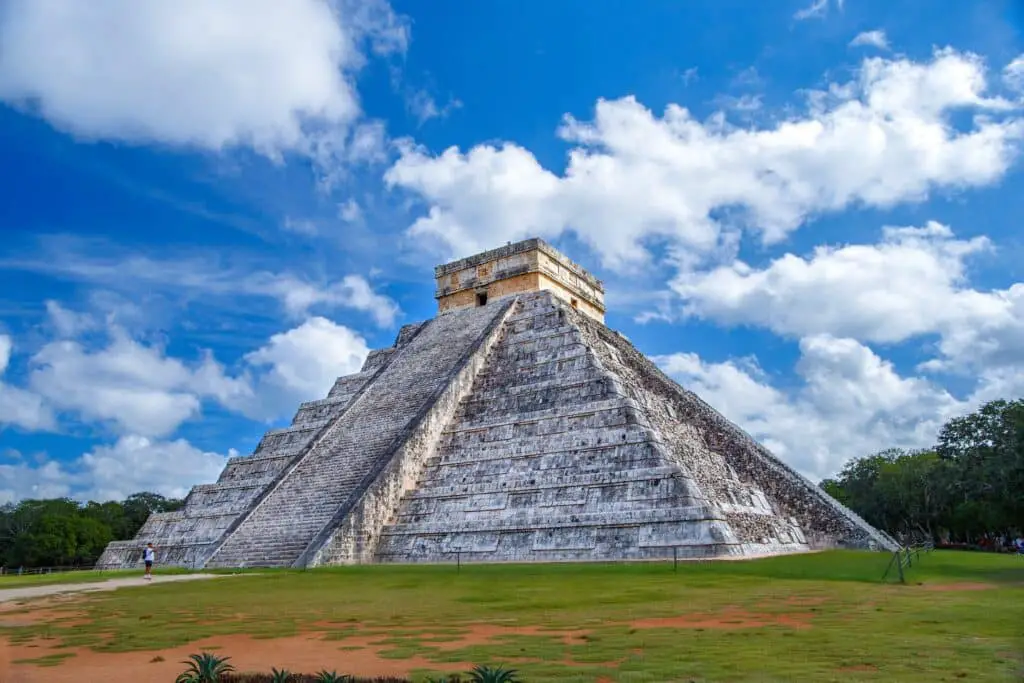 Large steep stone pyramid with stepped sides leading to a small doorway at the top, which has a flat roof. The sky is blue with fluffy white clouds and green grass around the pyramid.