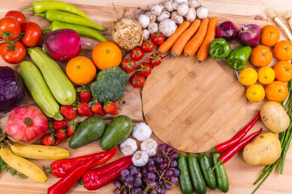 Looking down at a wooden table full of colorful whole fruits and vegetables - green zucchinis and peppers, white garlic bulbs, oranges and carrots, bunches of purple grapes, red chili peppers, tomatoes and strawberries. 