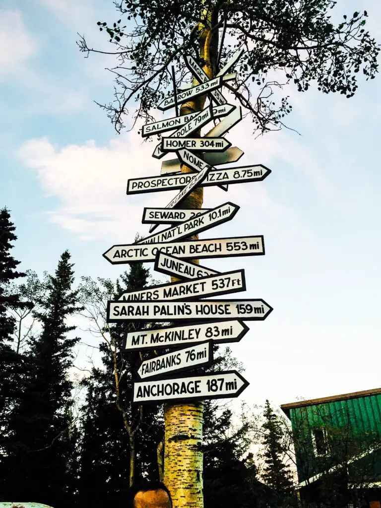 Tall sign in Alaska with arrows with names of points of interest how many miles places in Alaska are from the sign, like Homer (304 mi) Fairbanks (76 mi), Sarah Palin's house (159 mi) and Prospector Pizza (8.75 mi).