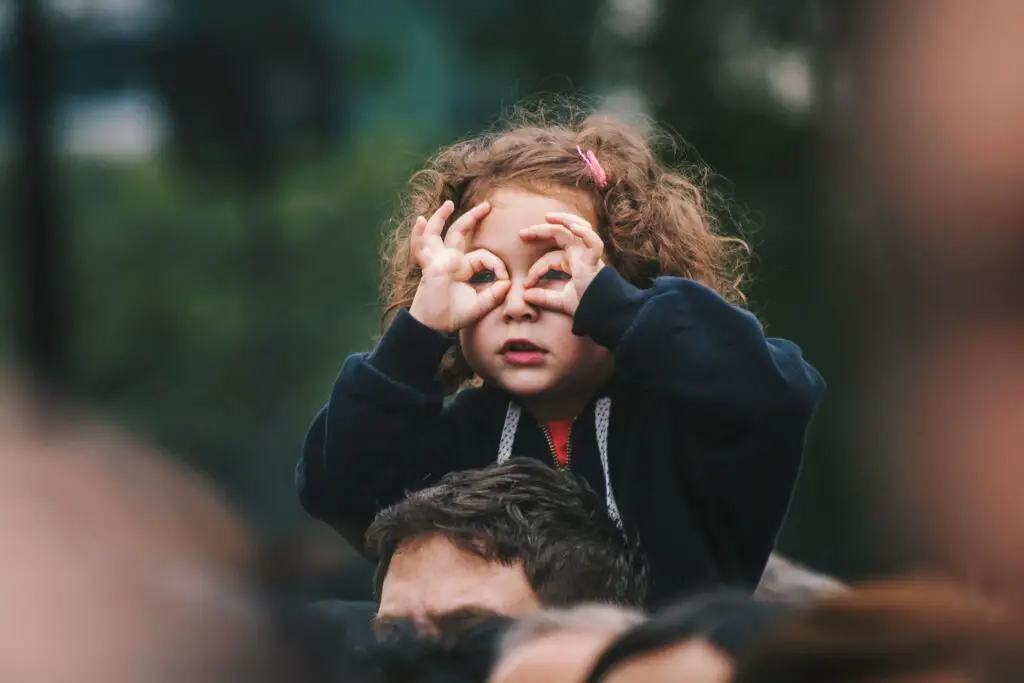 Little girl sitting on a man's shoulders holding her hands up to her eyes encircling her eyes with her fingers like pretend binoculars.