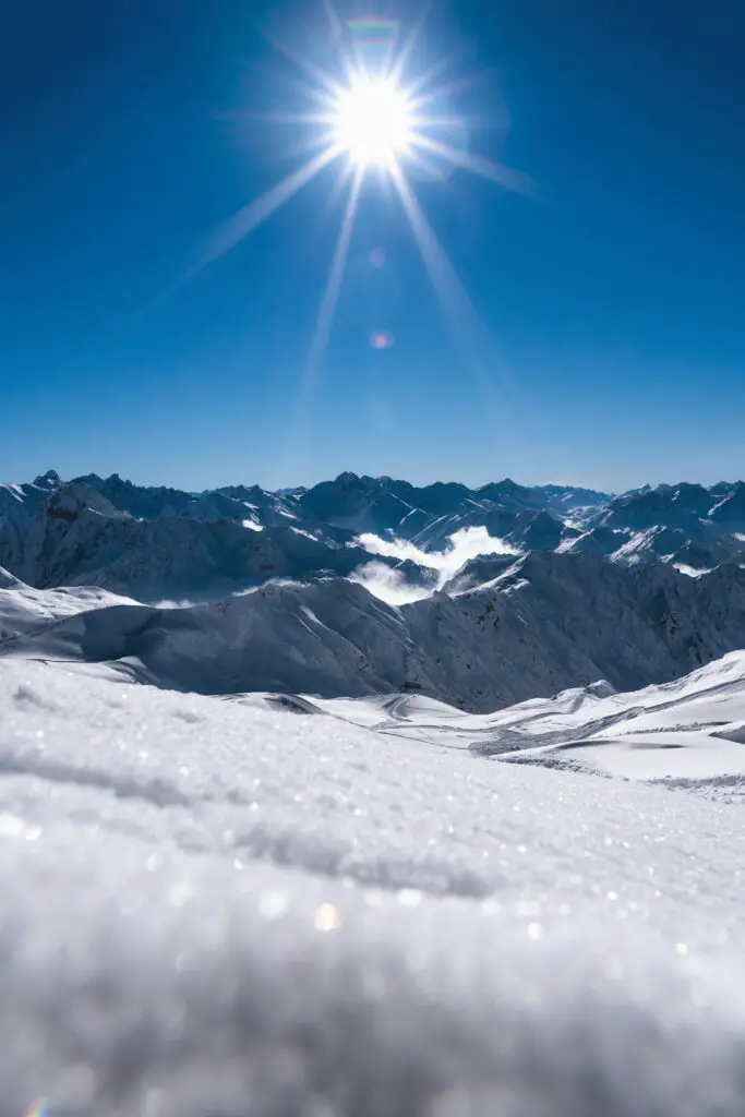 Snow-covered ground with grey, rocky mountains in the background. A large bright sun shines in a blue sky. The sun reflecting on the snow is very bright.