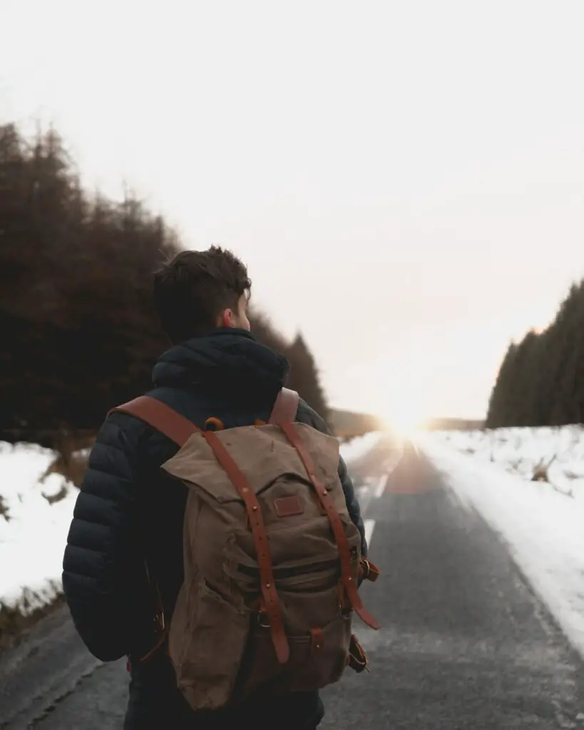 Man standing facing away from the camera on a snow-lined road. He is wearing a green canvas backpack.