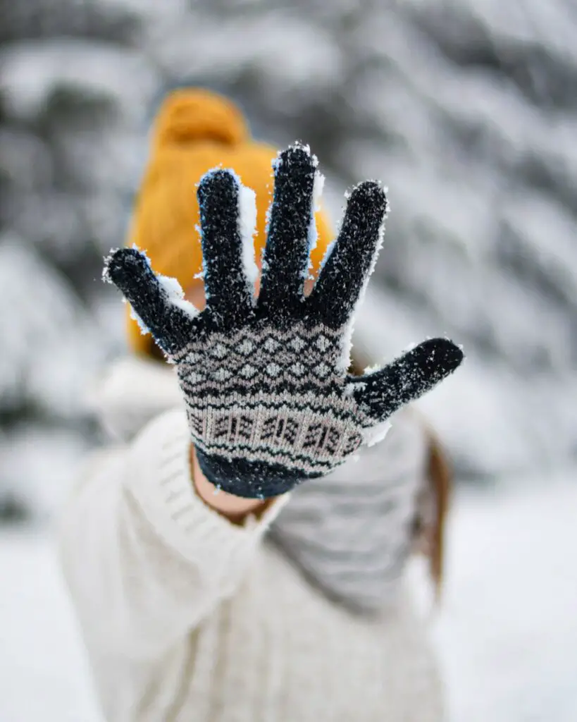 Girl wearing a white coat and mustard-yellow hat holding her knit gloved-covered palm, with fingers spread, up to the camera, hiding her face.
