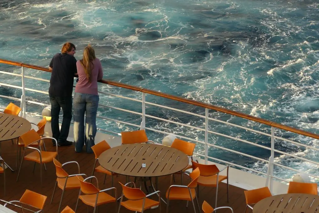 A man and a woman leaning on the railing of a cruise ship deck looking out over the water