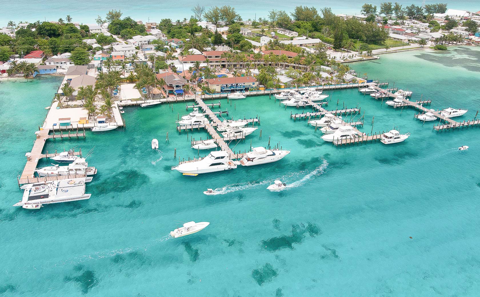 Aerial view of boat marina on a narrow strip of land with small buildings, palm trees and white sand beaches with crystal clear turquoise ocean