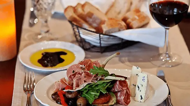 In the background, a basket of bread sits beside a shallow dish of olive oil and balsamic vinegar. A plate of antipasti is in the foreground with meat, cheese, greens and olives