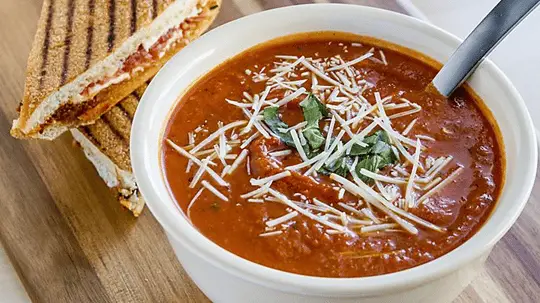 White bowl of tomato-looking soup topped with shredded cheese and cilantro, with a grilled sandwich on the side.