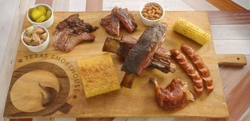 Platter of BBQ'd foods - chicken ribs, brisket with a sides of cornbread, beans, corn on the cob, pickles and potato salad.