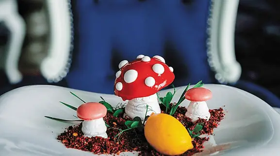 A white plate of food formed to look like cartoon mushrooms with white bases and red caps, sitting in a bed of something made to look like dirt with tufts of "grass." A lemon wedge sits on the edge.