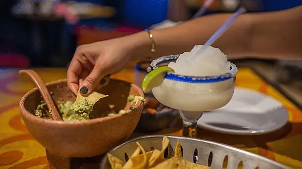Woman's hand dipping a tortilla chip into a wooden bowl of guacamole which sits beside a frosty blended beverage that looks like a pina colada