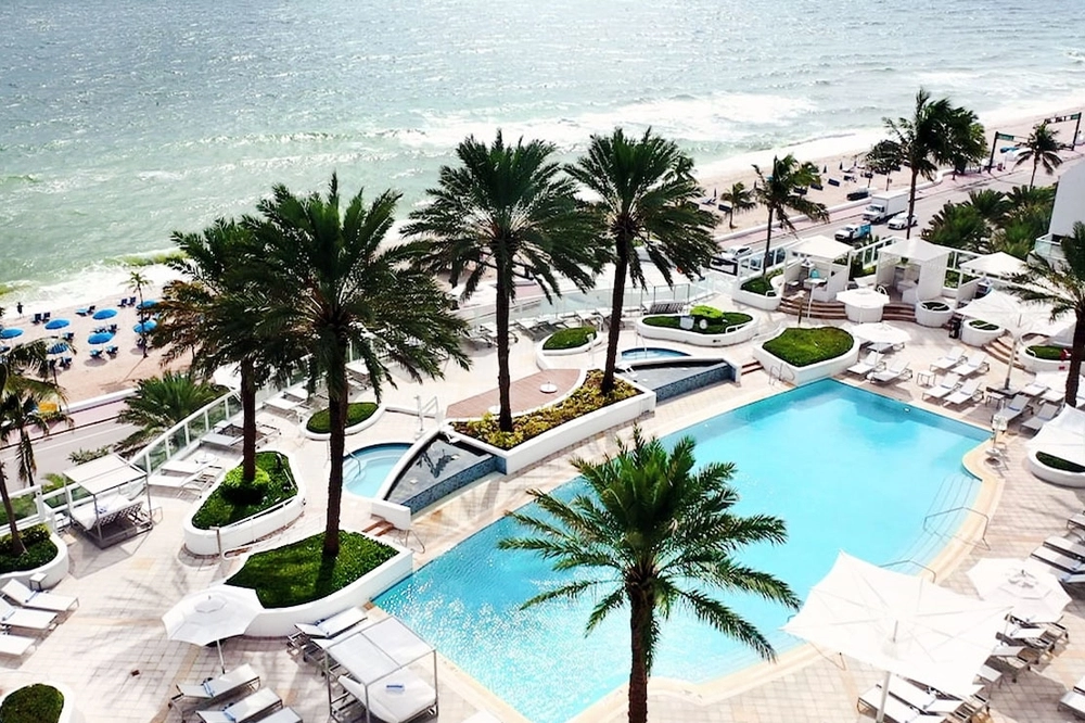Aerial view of pool surrounded by palm trees adjacent to the beach and ocean at the Hilton Fort Lauderdale Beach Resort.