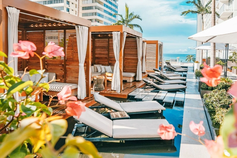 Line of wooden cabanas with privacy curtains pulled back with loungers sitting in shallow pools. The beach and ocean is seen in the background. Alohilani Resort Waikiki Beach.