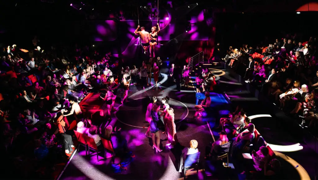 Performance in The Red Room. The cast is up close to the guests sitting is small groups around tables. The room is dark and lit with colors of blue and purple.