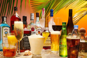 Against a tropical-looking background, there are bottles of different types of alcohol, beers and wine, plus glasses of beer, champagne and cocktails.