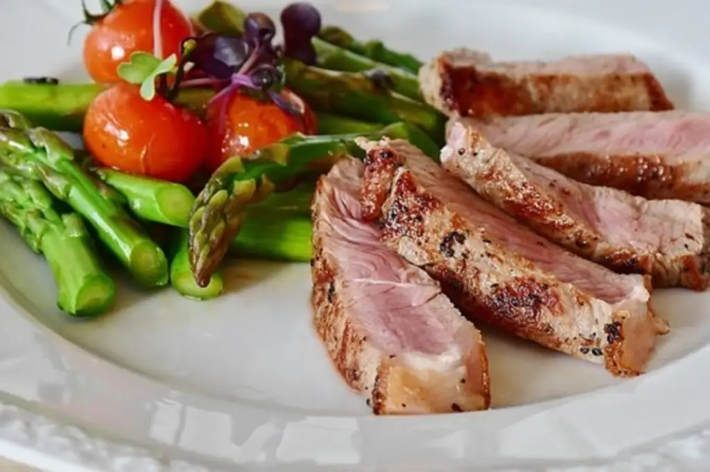 White plate of keto-friendly foods - sliced medium-cooked steak, a pile of asparagus topped with seared cherry tomatoes.
