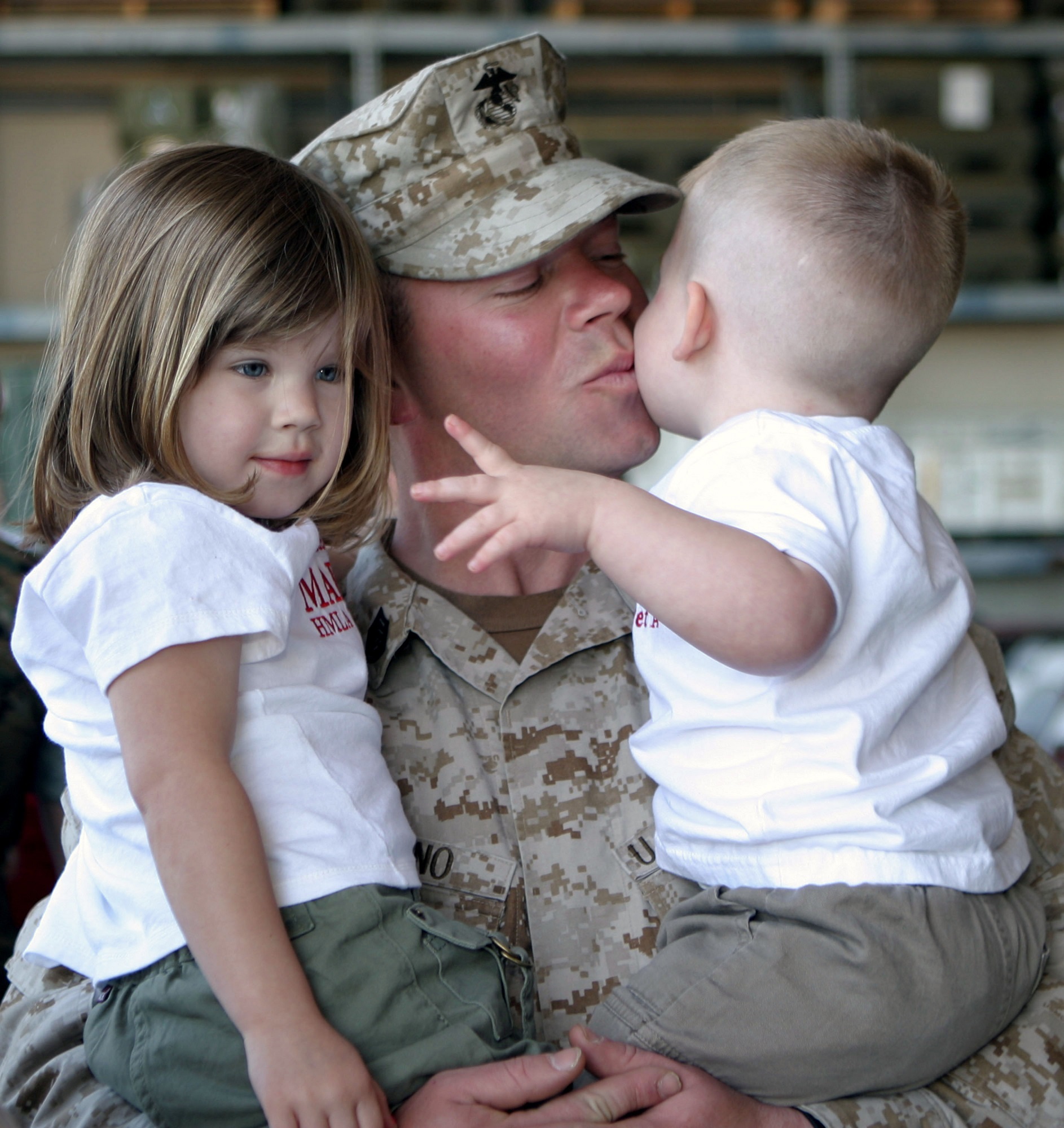 Man dressed in army camouflage holding two young children, and kissing one child on the cheek.