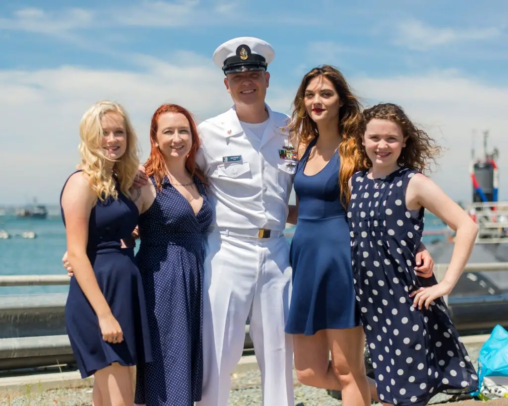 Family with arms around each other smiling at the camera. The man is dressed in a white military uniform and the women (presumably wife and 3 teen girls) wearing dresses. They look to be standing on the deck of a ship.