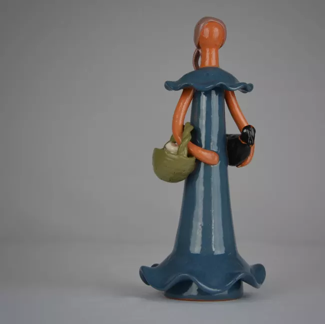 Ceramic "doll" with blue dress, no face, long hair pulled back, carrying a basket in on hand and black duck in the other