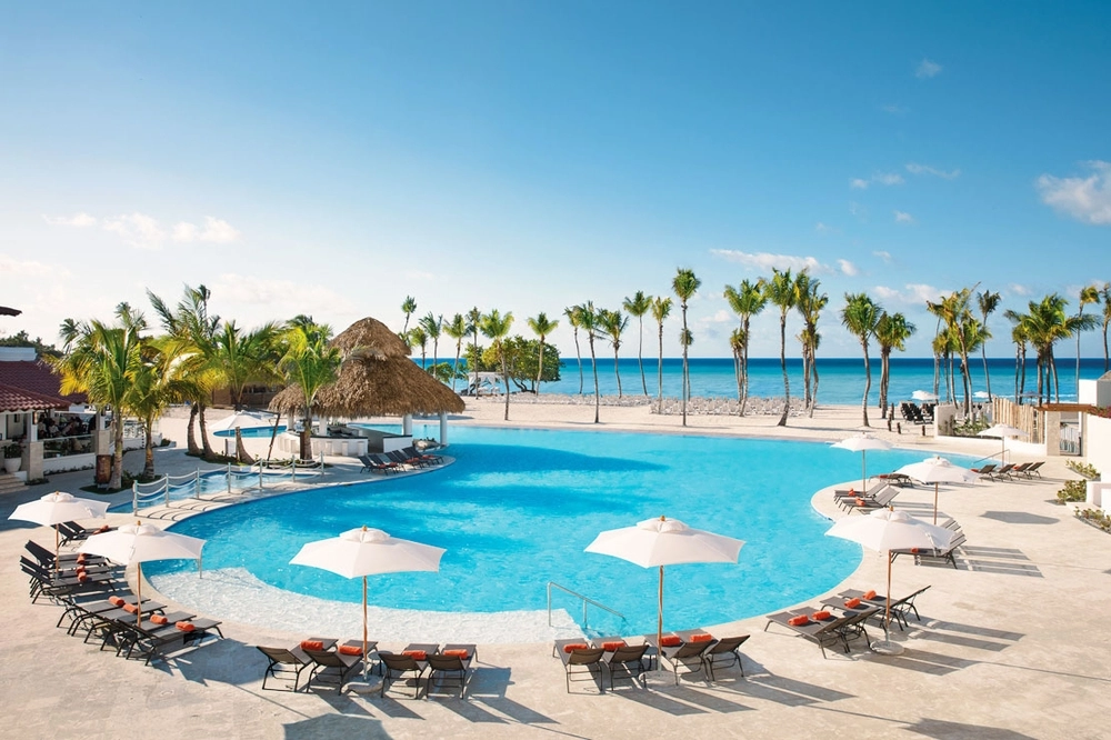 View of large resort pool surrounded by loungers and white umbrellas. Just beyond the pool is the white sand beach lined with skinny palm trees and turquoise water.
