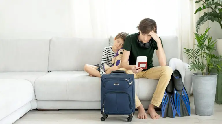 A sad looking boy and man sitting on a couch holding tickets and passports in hand and a suitcase stands in front of them.